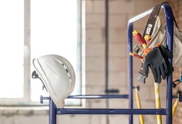 White construction hard hat and tool belt with gloves hanging on blue scaffolding at a job site.