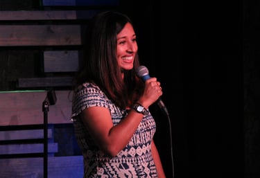 A woman smiles on stage with a microphone, dressed in a patterned dress, in a dimly lit comedy venue.