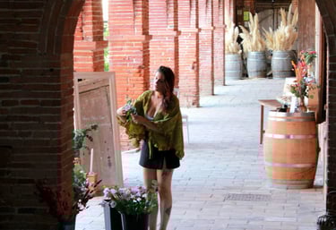 Estelle arrangeant des fleurs séchées dans des seaux sous une galerie en briques avec des arches