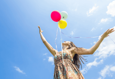 Mujer joven en un espacio abierto que muestra un cielo azul, lanzando tres globos al aire, feliz.