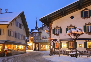 Snowy Bavarian village street at dusk with illuminated traditional alpine chalets and shops.