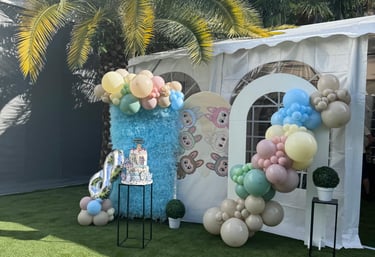 Outdoor birthday party decoration with a balloon arch, tiered cake, and palm tree on a green lawn.