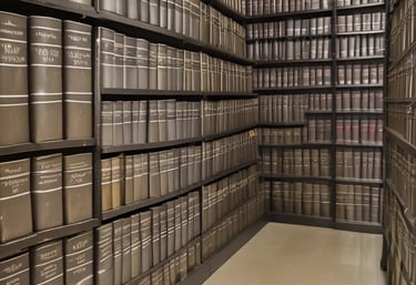 A person in a suit is seated at a desk, holding a phone to their ear and using the other hand to turn pages in a document. The workspace includes a computer monitor, paperwork, and other office supplies. Large windows allow natural light to illuminate the area.