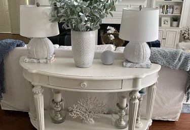 Painted sofa table in off white with light distressing with two lamps and a vase with greenery. 