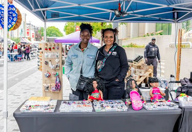 Two women smiling at an outdoor Afro Fiesta Festival market stall selling handmade accessories and dolls.