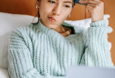 A thoughtful woman holding a credit card while shopping online with a laptop in bed.