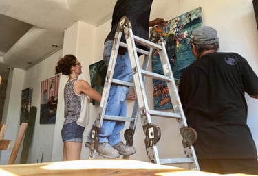 Professional art installers using a ladder to hang colorful paintings in a modern gallery.