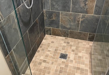 Walk-in shower with dark slate wall tiles, tan mosaic floor, and chrome fixtures.