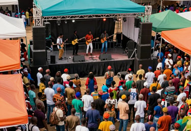 An overhead view of a lively African / Pan-African outdoor event. Bright teal and orange canopies provide shade for a crowd gathered around a central stage where a music group is performing.
