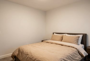 A professional real-estate photograph of a cozy and clean master bedroom. The walls are warm off-white and the bedding is soft tan, creating a welcoming and dependable aesthetic.