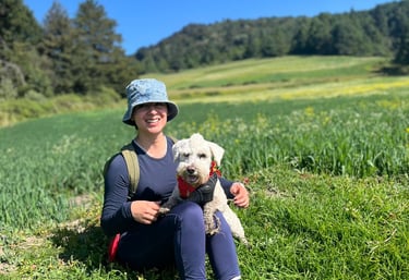 Una mujer sonriente está sentada en un prado verde con su perro blanco mientras hacen senderismo