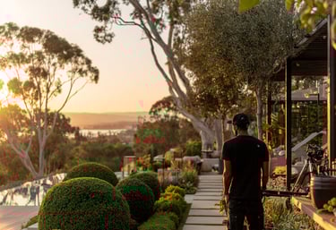 A photographer stands on a luxury patio with manicured gardens overlooking a sunset ocean view.