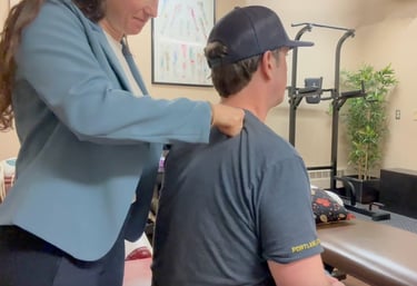 Chiropractor performing deep tissue work on seated patient's shoulder in treatment room. 
