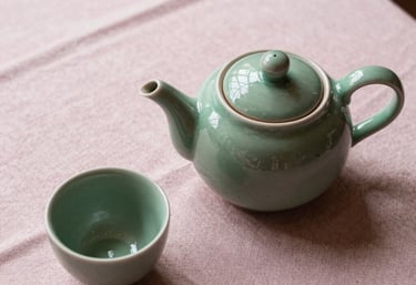 A top-down view of a ceramic teapot and a single sage green cup on a linen tablecloth, soft pearl pink light, Southern European / Italian morning.