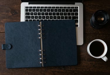 A top-down view of a minimalist North American / US workspace with a laptop, a dark navy planner, and a cup of coffee.
