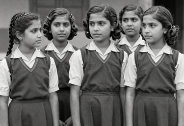 A nostalgic black and white photograph of South Asian Pakistani girls in school uniforms from the 1960s standing in a row, soft vintage grain.
