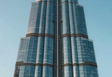 A close-up of a modern glass skyscraper in Dubai reflecting the soft blue and light cyan sky. The architecture is sharp and innovative.
