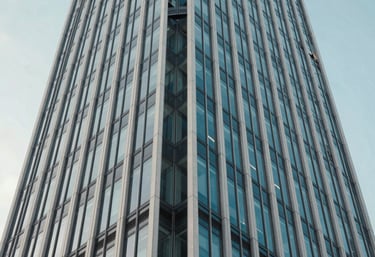 An abstract, low-angle photograph of a modern skyscraper's steel and glass facade against a light blue sky. The composition conveys strength, stability, and a future-forward vision. Muted blue and off-white color tones.