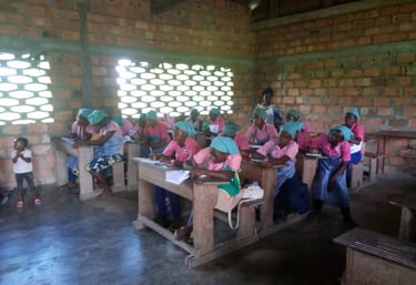 Students in uniforms writing at wooden desks inside a brick classroom for community education.