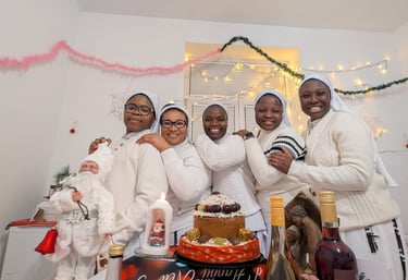 Smiling group of five nuns in white habits celebrating Christmas with a cake, wine, and festive decorations.