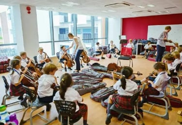 Students in school uniforms practice playing cellos and violins during a music class in a bright classroom.