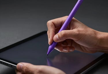 A close-up of a designer's hands using a sleek black tablet with a purple stylus in a dark, minimalist studio. North American / International.