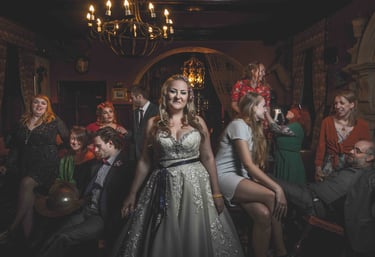 Smiling bride in a lace wedding gown at a vintage style reception with guests and chandelier.