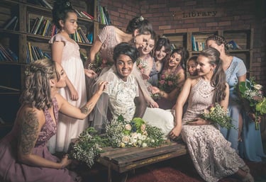 Smiling bride in a white gown surrounded by bridesmaids in pink and blue dresses in a rustic library setting.