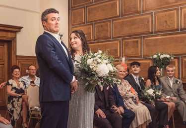 A groom making a funny face during a wedding ceremony in a wood-paneled room while the bride looks on.