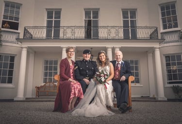 A bride and groom in military uniform sit with family outside a white manor wedding venue.