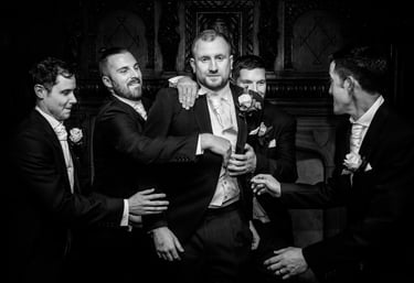 Black and white photo of a groom and four groomsmen in formal wedding suits adjusting his jacket.