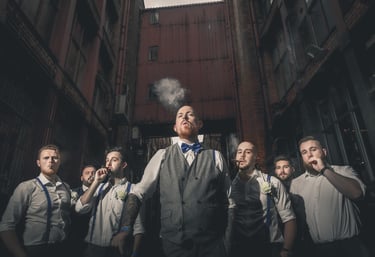 Groom and groomsmen in vintage wedding attire smoking cigars in an industrial alleyway.