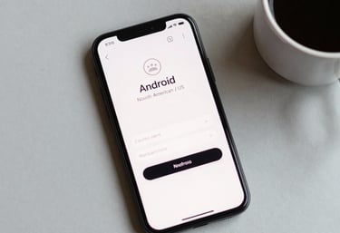 A top-down photograph of a minimalist Android smartphone and a coffee cup on a light grey desk. The screen shows a clean login interface. Soft North American / US office lighting.