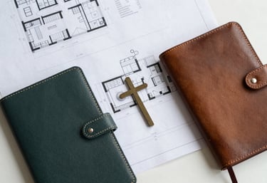 An overhead shot of a clean workspace featuring architectural plans, a leather journal, and a small cross, symbolizing strategic planning and integrity. The palette uses professional tones of #3D5B5F and #A2B2AE, reflecting dignified stewardship and accountability.