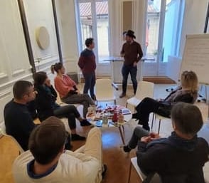 Olivier Paris interrogeant un participant tout les deux debout devant six autres participants assis