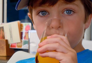 A young boy with striking blue eyes drinks a glass of orange juice at an outdoor cafe.