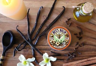 All Natural Vanilla Spice Bee's Balm tin surrounded by vanilla beans, cloves, olive oil, and candle