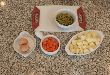 a cutting board on a counter with eggs, potatoes, carrots and peas