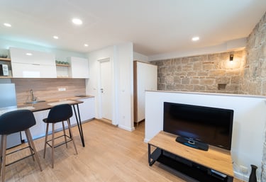 living room with walls made of stone, tv and white fully equiped kitchen