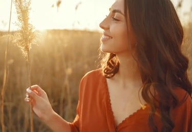 Une femme souriante se tient dans un champ, dans la lumière du soleil couchant, évoquant la simplicité et la confiance.