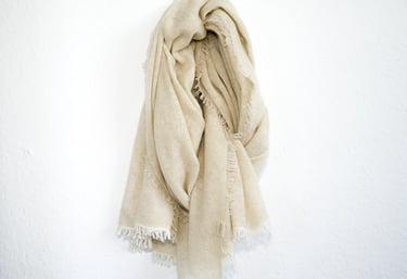 Cream pashmina shawl with fringed edges hanging on white wall, soft texture and cozy appearance.
