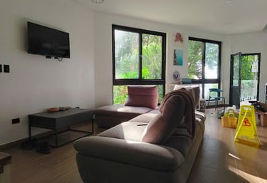 Modern bright living room with a large plush sectional sofa, wall-mounted TV, and floor-to-ceiling windows overlooking lush