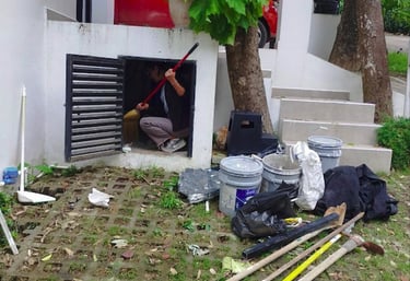 A worker cleaning a small outdoor storage unit with a broom near gardening tools and buckets.