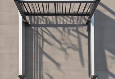 A wooden pergola structure extends over a concrete walkway with tall buildings in the background and clear blue skies above. Metal lampposts are positioned along the path, providing lighting. Trees and greenery can be seen on the left side of the image, adding a touch of nature to the urban setting.