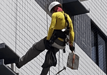 Técnico alpinista realizando teste de percussão e remoção de entulho em fachada predial, durante ins