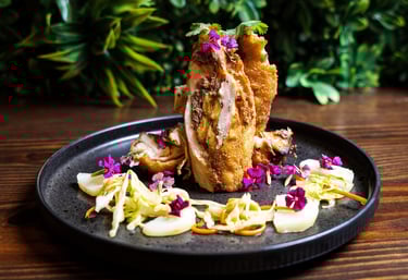 Gourmet stuffed chicken rollatini with plantains, garnished with edible purple flowers and slaw on a black plate.