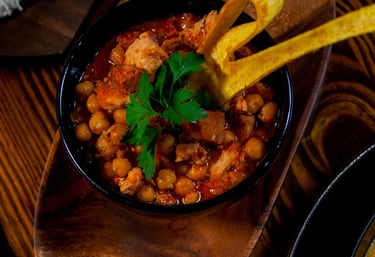 A bowl of Spanish chickpea stew with pork and plantain chips on a rustic wooden tray.