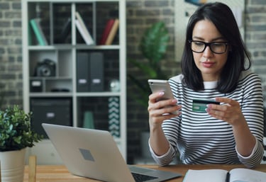 Eine Frau hat ihre Bankkarte in der Hand, sitzt vor einem Schreibtisch mit ihrem Laptop darauf.