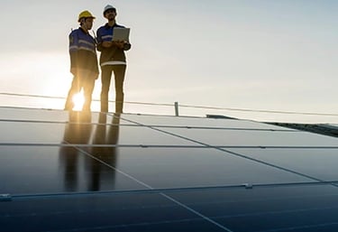 Two solar technicians in hard hats and safety gear inspecting rooftop solar panels with a laptop.