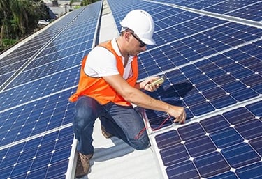 A solar panel installer in a safety vest and hard hat testing blue photovoltaic panels on a roof.